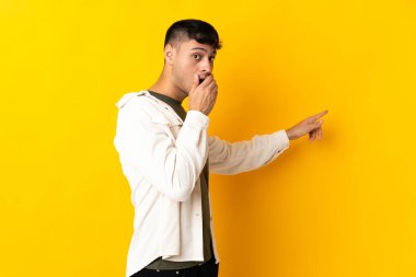 Young Colombian man isolated on yellow background with surprise expression while pointing side clipart