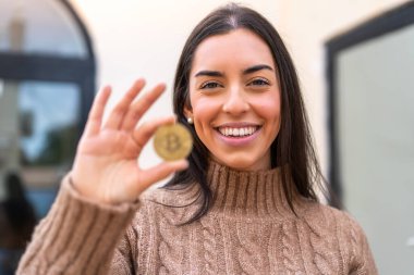 Açık havada elinde Bitcoin tutan genç bir kadın mutlu bir ifadeyle