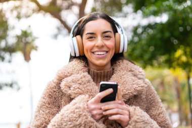 Genç esmer kadın açık havada cep telefonuyla müzik dinliyor ve ön tarafa bakıyor.