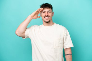 Beyaz tenli yakışıklı adam mavi arka planda izole edilmiş mutlu bir ifadeyle selamlıyor.