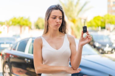 Genç ve güzel bir kadın dışarıda arabanın anahtarlarını tutuyor, üzgün bir ifadeyle.