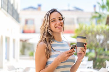 Açık havada cep telefonu kullanan ve elinde mutlu bir ifadeyle kahve tutan genç ve güzel bir kadın.