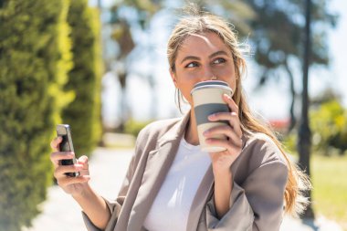 Genç güzel sarışın Uruguaylı kadın açık havada cep telefonu kullanıyor ve elinde kahve tutuyor.