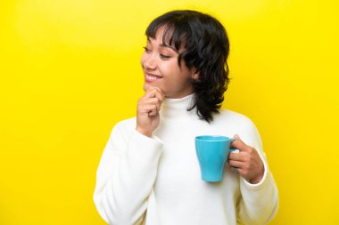 Arjantinli genç bir kadın elinde kahve fincanıyla sarı arka planda izole edilmiş bir şekilde bir fikir üretiyor ve bakıyor.