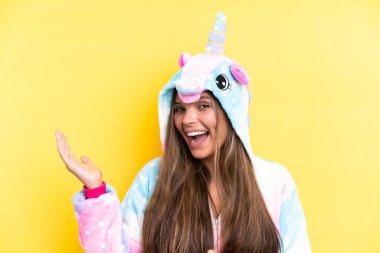 Tek boynuzlu bir pijama giyen genç beyaz kadın sarı arka planda izole edilmiş ve davet edilmek için ellerini yana uzatıyor.