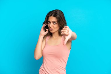 Mavi arka planda izole edilmiş cep telefonu kullanan genç beyaz kadın olumsuz ifade ile baş parmağını gösteriyor.