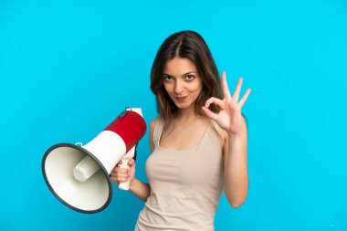 Mavi arka planda izole edilmiş genç beyaz bir kadın elinde bir megafonla tamam işaretini parmaklarıyla gösteriyor.