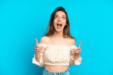 Mavi arka planda izole edilmiş genç beyaz kadın şaşırmış ve yukarı bakıyor.