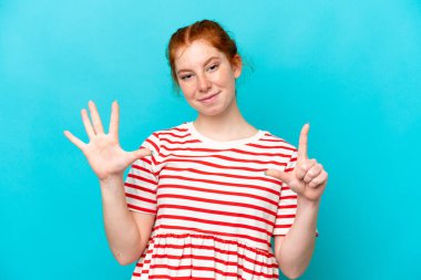 Mavi arka planda izole edilmiş genç kızılımsı kadın parmaklarıyla yediyi sayıyor