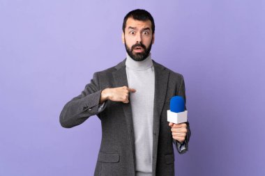 İzole edilmiş mor arka plan üzerinde mikrofon tutan sakallı yetişkin bir muhabir kendini işaret ediyor.