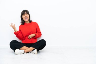 Genç melez kadın, beyaz arka planda izole bir şekilde yerde oturmuş gitar hareketi yapıyor.