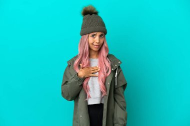Pembe saçlı, yağmura dayanıklı bir ceket giyen, mavi arka planda izole edilmiş genç bir kadın kendini işaret ediyor.