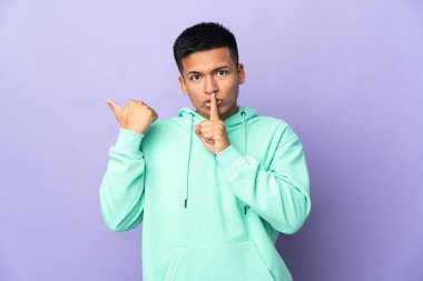 Mor arka planda izole edilmiş genç Ekvadorlu adam yan tarafı işaret ediyor ve sessizlik hareketi yapıyor.