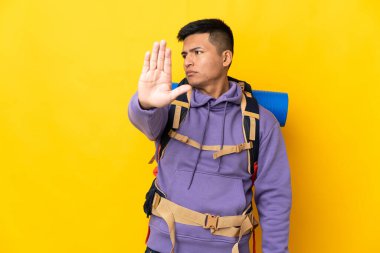 Young mountaineer man with a big backpack isolated on yellow background making stop gesture and disappointed