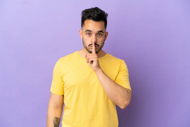 Mor arka planda izole edilmiş genç beyaz adam sessiz bir jest işareti gösteriyor. Parmağını ağzına sokuyor.