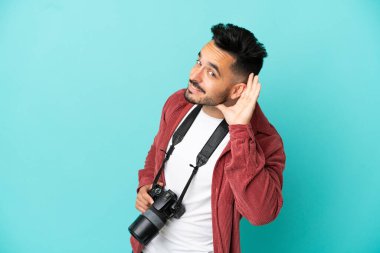 Genç fotoğrafçı, mavi arka planda izole edilmiş bir şekilde elini kulağına koyarak bir şeyler dinliyor.