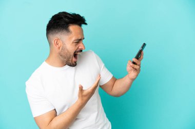 Genç beyaz adam mavi arka planda izole edilmiş cep telefonu kullanıyor ve şarkı söylüyor.
