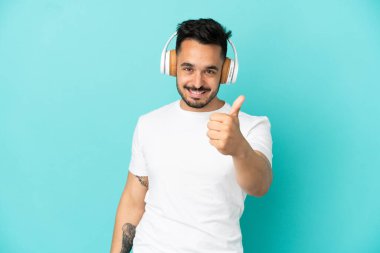 Kafkasyalı genç adam mavi arka planda izole edilmiş müzik dinliyor ve başparmağını kaldırıyor.