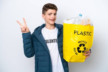 Beyaz arka planda geri dönüşüm yapmak için elinde plastik şişelerle dolu bir çanta tutan Rus ergen gülümseyerek zafer işareti gösteriyor.