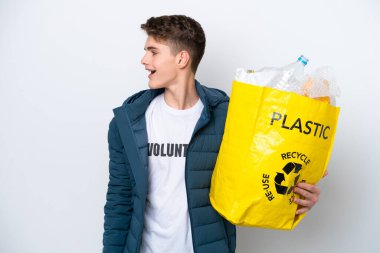 Beyaz arka planda geri dönüşüm yapmak için elinde plastik şişelerle dolu bir çanta tutan genç Rus. Yan pozisyonda gülerek.