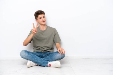 Genç Rus adam beyaz arka planda izole bir şekilde yerde oturuyor gülümsüyor ve zafer işareti gösteriyor.