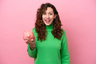 Young caucasian woman holding piggybank isolated on pink background with surprise and shocked facial expression