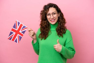 İngiliz bayrağını tutan beyaz bir kadın pembe arka planda izole edilmiş. Başparmakları havada. Çünkü iyi bir şey oldu.