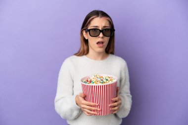 Mavi arka planda soyutlanmış beyaz bir kadın 3D gözlüklü ve elinde koca bir kova patlamış mısırla şaşırdı.