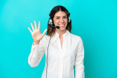Tele pazarlamacı İtalyan kadın mavi arka planda izole edilmiş kulaklıkla çalışıyor. Parmaklarıyla beşi sayıyor.