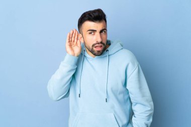 İzole edilmiş mavi arka planda beyaz bir adam. Kulağına elini sürerek bir şeyler dinliyor.