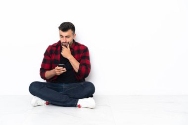 Young handsome man sitting on the floor thinking and sending a message