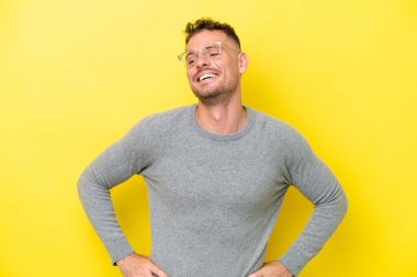 Sarı arka planda izole edilmiş, kolları kalçasında ve gülümseyen genç beyaz bir adam.