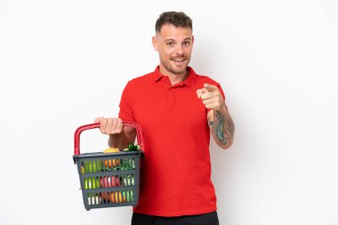 Beyaz arka planda izole edilmiş yiyecek dolu bir sepet taşıyan beyaz bir adam şaşırmış ve ön tarafı işaret ediyor.