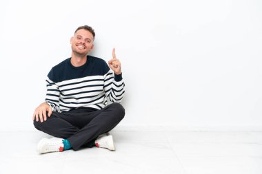 Genç adam beyaz arka planda izole bir şekilde yerde oturuyor ve en iyinin işareti olarak parmağını kaldırıyor.
