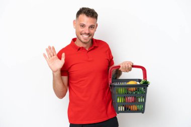Beyaz arka planda izole edilmiş, elinde yiyecek dolu bir sepet tutan beyaz bir adam mutlu bir ifadeyle selamlıyor.