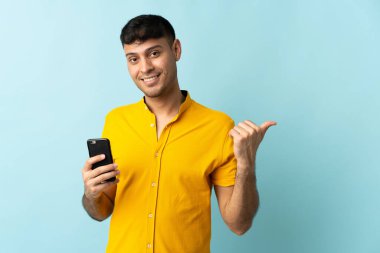 Cep telefonu kullanan genç Kolombiyalı bir adam mavi arka planda izole edilmiş bir ürün sunmak için kenarı işaret ediyor
