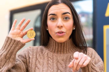 Elinde Bitcoin tutan genç bir kadın şaşırmış ve ön tarafı işaret ediyor.