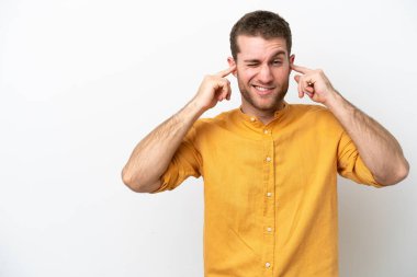 Beyaz arka planda izole edilmiş genç beyaz adam hayal kırıklığına uğramış ve kulaklarını kapatmış.