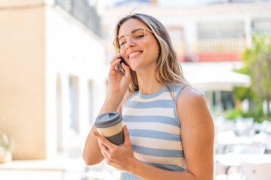 Açık havada cep telefonu kullanan ve elinde mutlu bir ifadeyle kahve tutan genç ve güzel bir kadın.