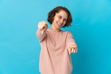 Mavi arka planda izole edilmiş genç bir İngiliz kadın mutlu bir ifadeyle ön tarafı işaret ediyor.