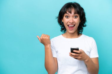 Genç Arjantinli kadın mavi arka planda izole edilmiş cep telefonu kullanıyor ve yan tarafı işaret ediyor.