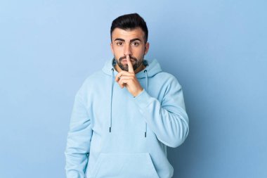 İzole edilmiş mavi arka planda beyaz bir adam sessizlik işareti gösteriyor. Parmağını ağzına sokuyor.