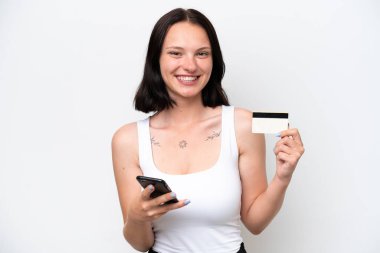 Beyaz arka planda izole edilmiş genç beyaz bir kadın cep telefonuyla kredi kartıyla alışveriş yapıyor.