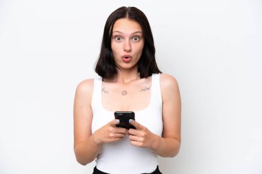 Beyaz arka planda izole edilmiş genç beyaz bir kadın kameraya bakarken şaşırmış bir ifadeyle cep telefonunu kullanıyor.