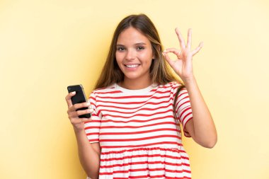 Sarı arka planda izole edilmiş genç beyaz kadın cep telefonu kullanıyor ve tamam işareti yapıyor.