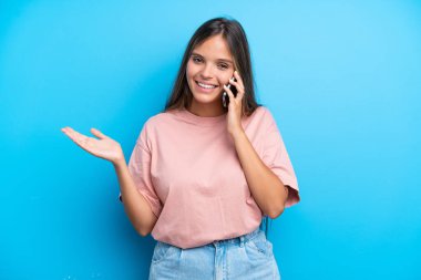 Mavi arka planda izole edilmiş genç beyaz bir kadın cep telefonuyla sohbet ediyor.