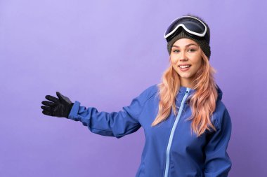 Kar kayağı gözlüklü genç kız izole edilmiş mor arka planda ellerini yana uzatarak davet ediyor.