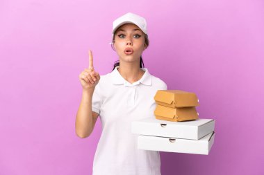 Pizzacı kız iş üniformalı pembe arka planda izole edilmiş pizza kutuları ve hamburgerleri topluyor. Çözümü fark etmek için parmağını kaldırıyor.