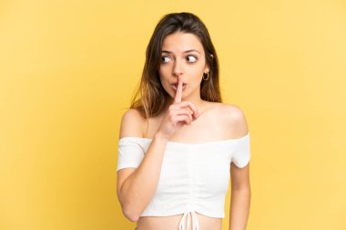 Sarı arka planda izole edilmiş genç beyaz bir kadın sessiz bir jest işareti gösteriyor. Parmağını ağzına sokuyor.
