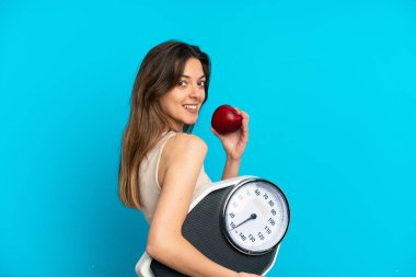 Mavi arka planda, tartı makinesi ve elmasıyla izole edilmiş genç beyaz bir kadın.